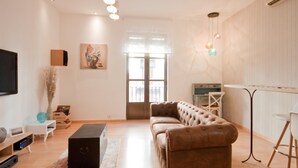 Un salón moderno con un sofá de cuero marrón, un reposapiés negro, una mesita auxiliar blanca y una mesa de centro de vidrio. Se aprecia una ventana grande con vistas al exterior, un cuadro en la pared y un televisor montado.