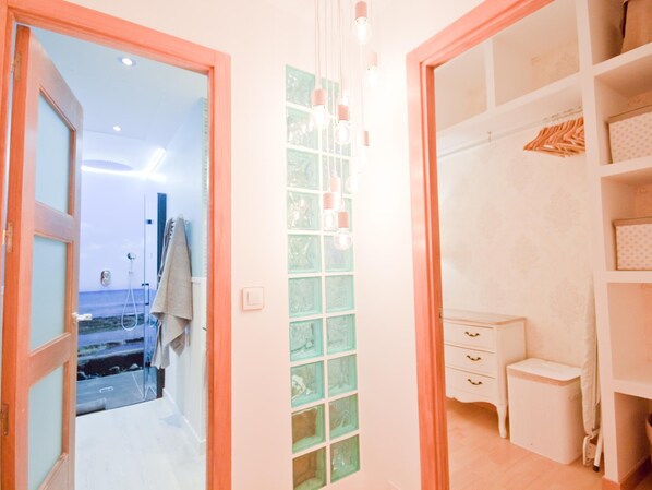 A hotel room with a view of the sea through a glass door, a small white bedside table, and a wardrobe with hanging clothes.