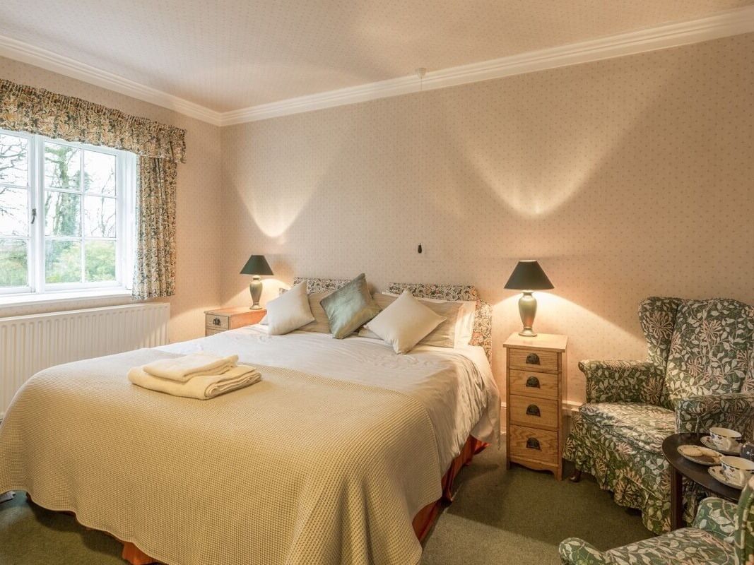A neatly arranged bedroom with a large bed, two bedside tables with lamps, a floral-patterned armchair, and a view of the outdoors through a window with curtains.
