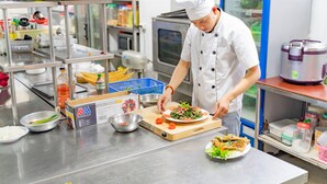 A chef preparing food in a commercial kitchen with stainless steel surfaces, various cooking utensils, and a refrigerator in the background. - Draco QK3 Cat Ba (Hai Phong)