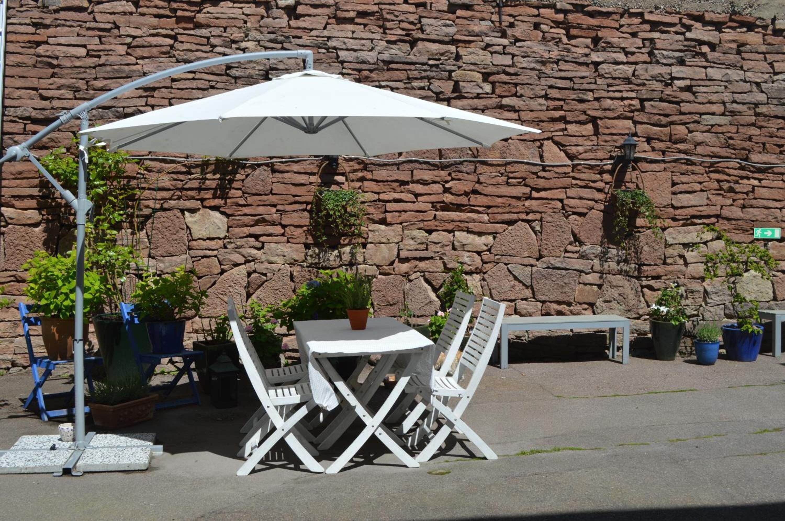 Un espacio al aire libre con mesa y sillas, una sombrilla y plantas en macetas, todo frente a un muro de piedra.