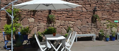 Un espacio al aire libre con mesa y sillas, una sombrilla y plantas en macetas, todo frente a un muro de piedra.