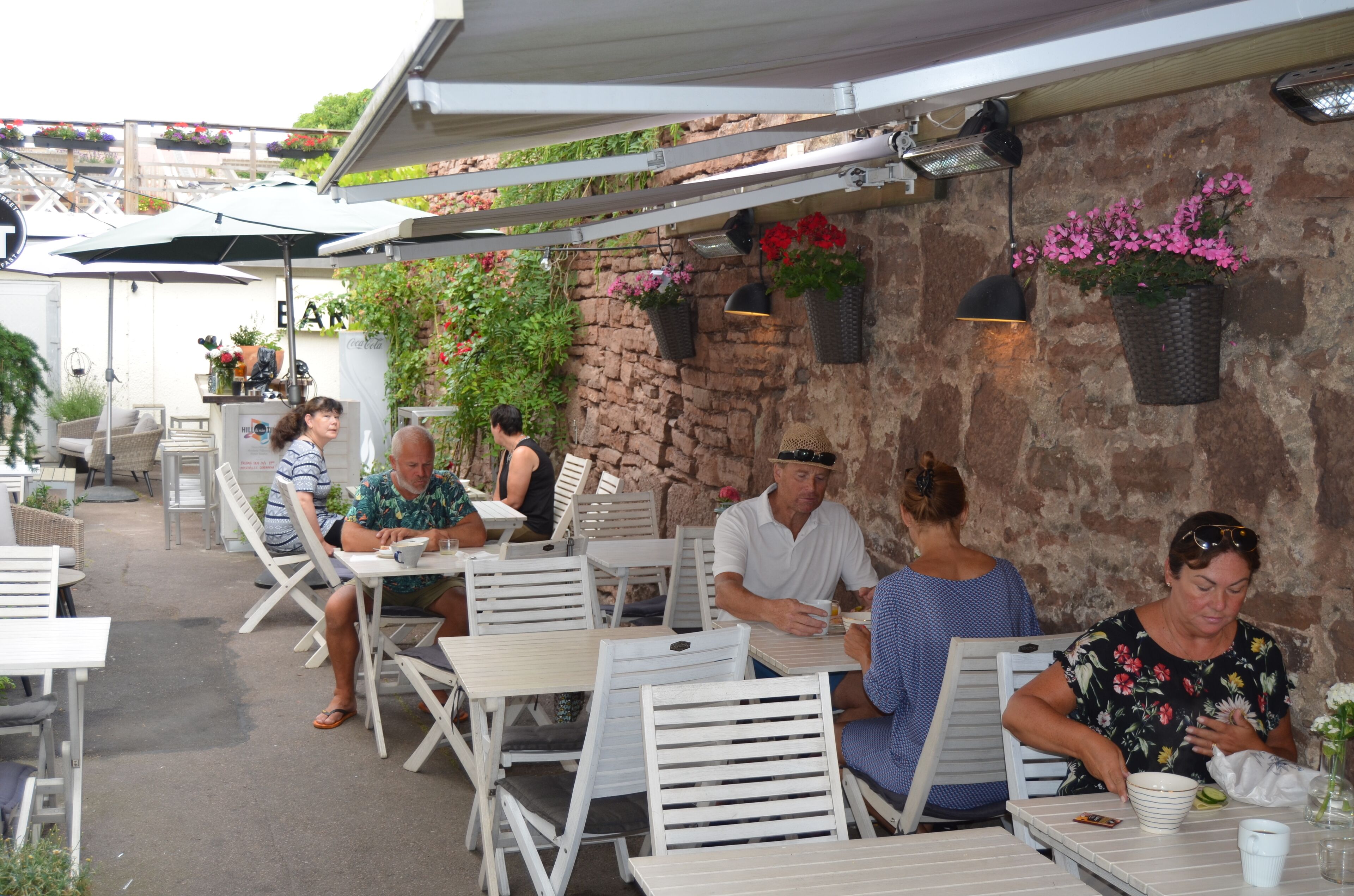 An outdoor dining area with white tables and chairs, a stone wall with hanging flower baskets, and a bar area in the background.