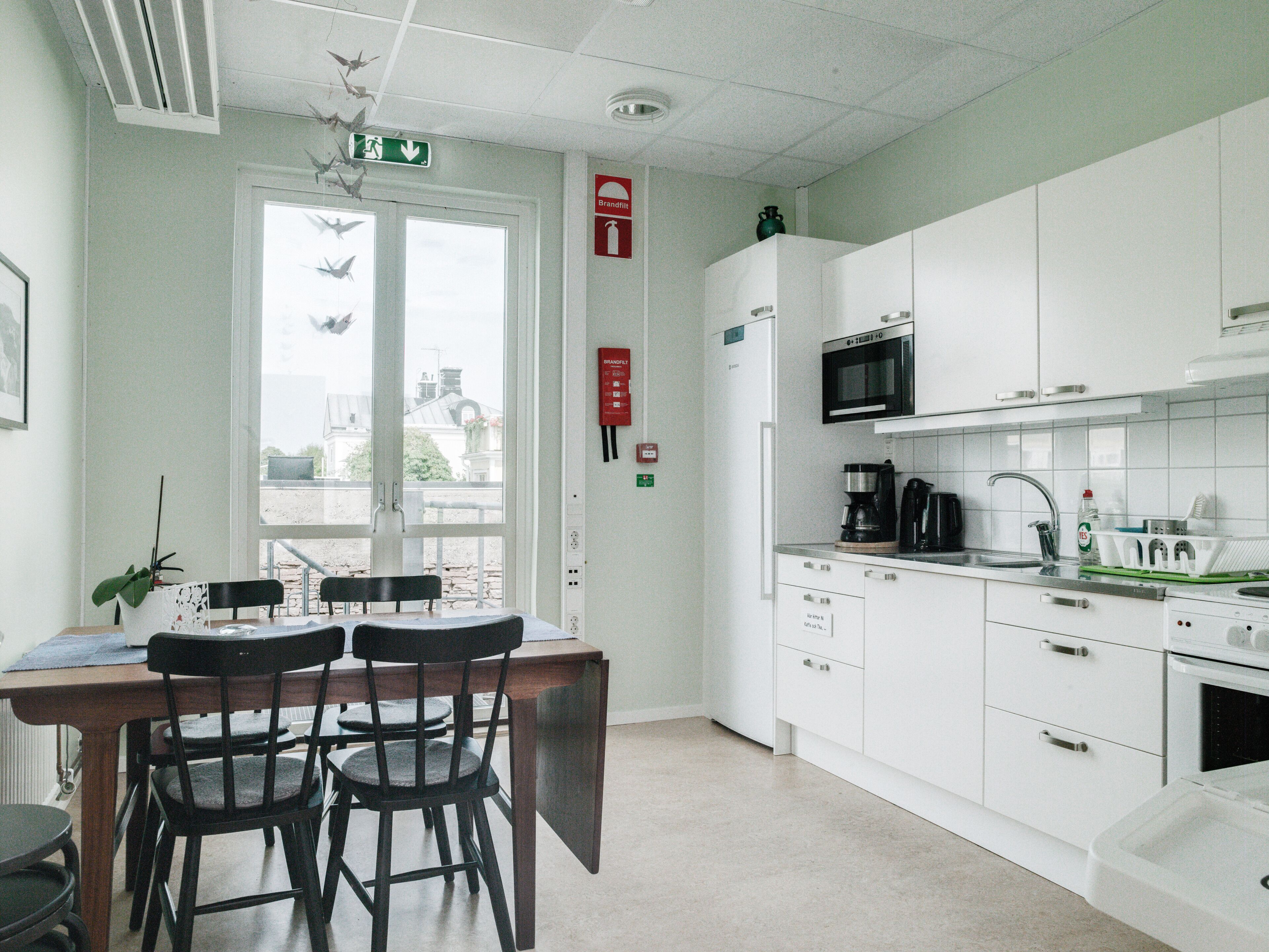 A modern kitchen with white cabinets, a microwave, and a coffee maker. There is a dining table with chairs and a window with a view of buildings outside.