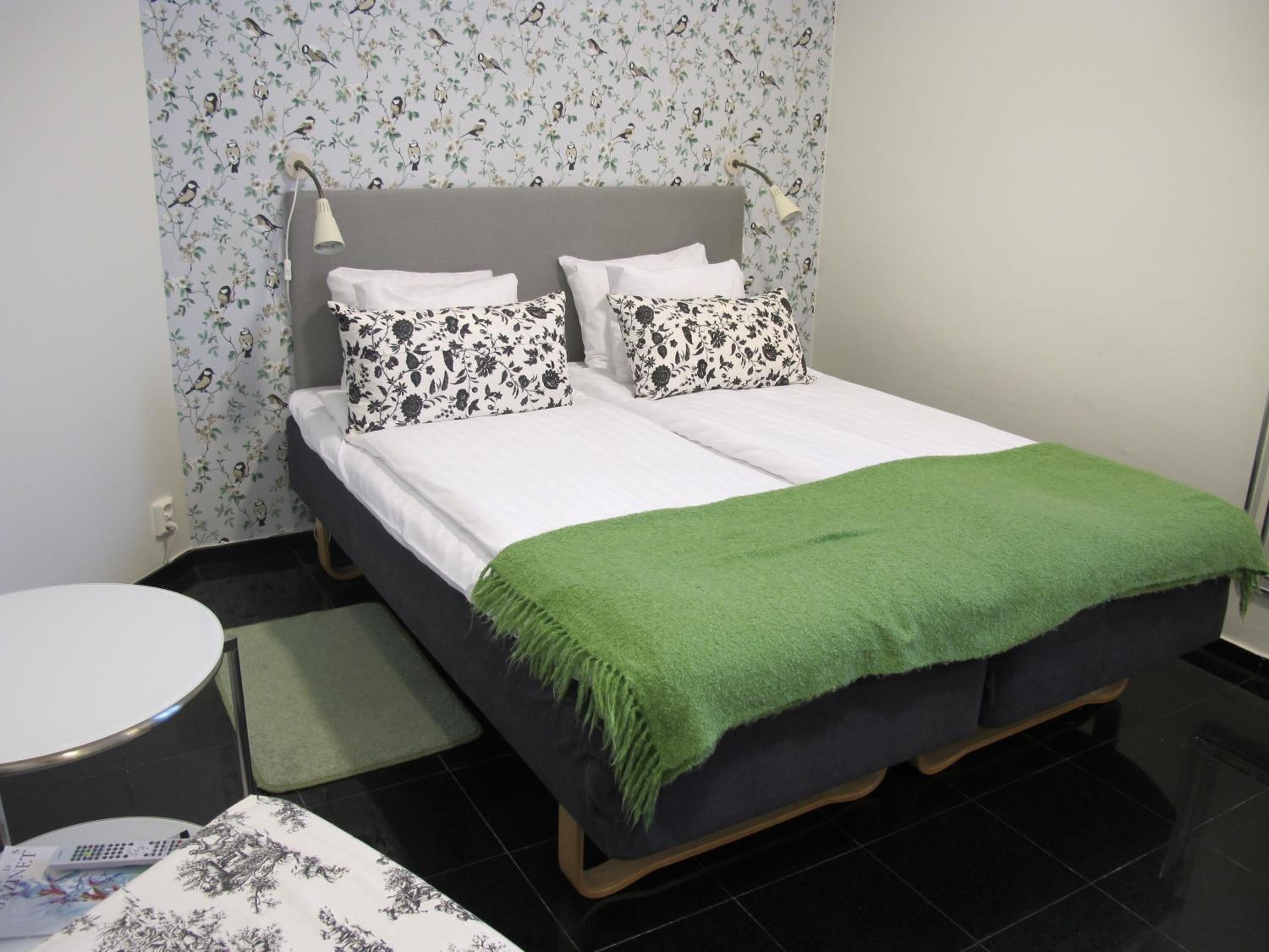 A neatly made double bed with a green throw blanket, two patterned pillows, and a grey headboard.