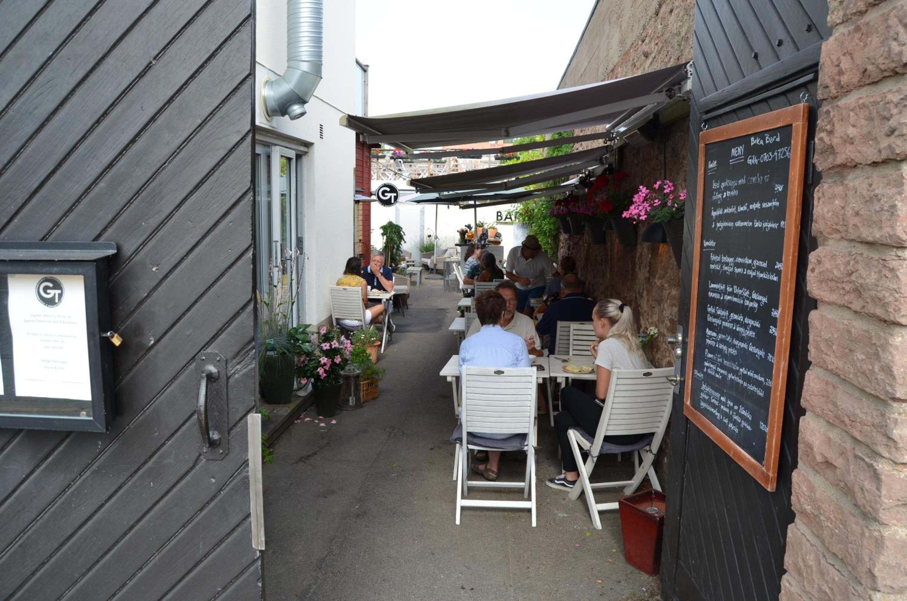An outdoor café with tables and chairs, a menu board, and a building with a 'GT' sign.