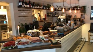 A bar area with a variety of bottles and glasses on shelves, a counter with food and drinks, and a kitchen visible in the background.