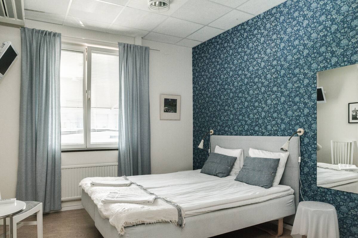 A bedroom with a bed, a bedside table, a small stool, a window with curtains, and a wall with a floral pattern.