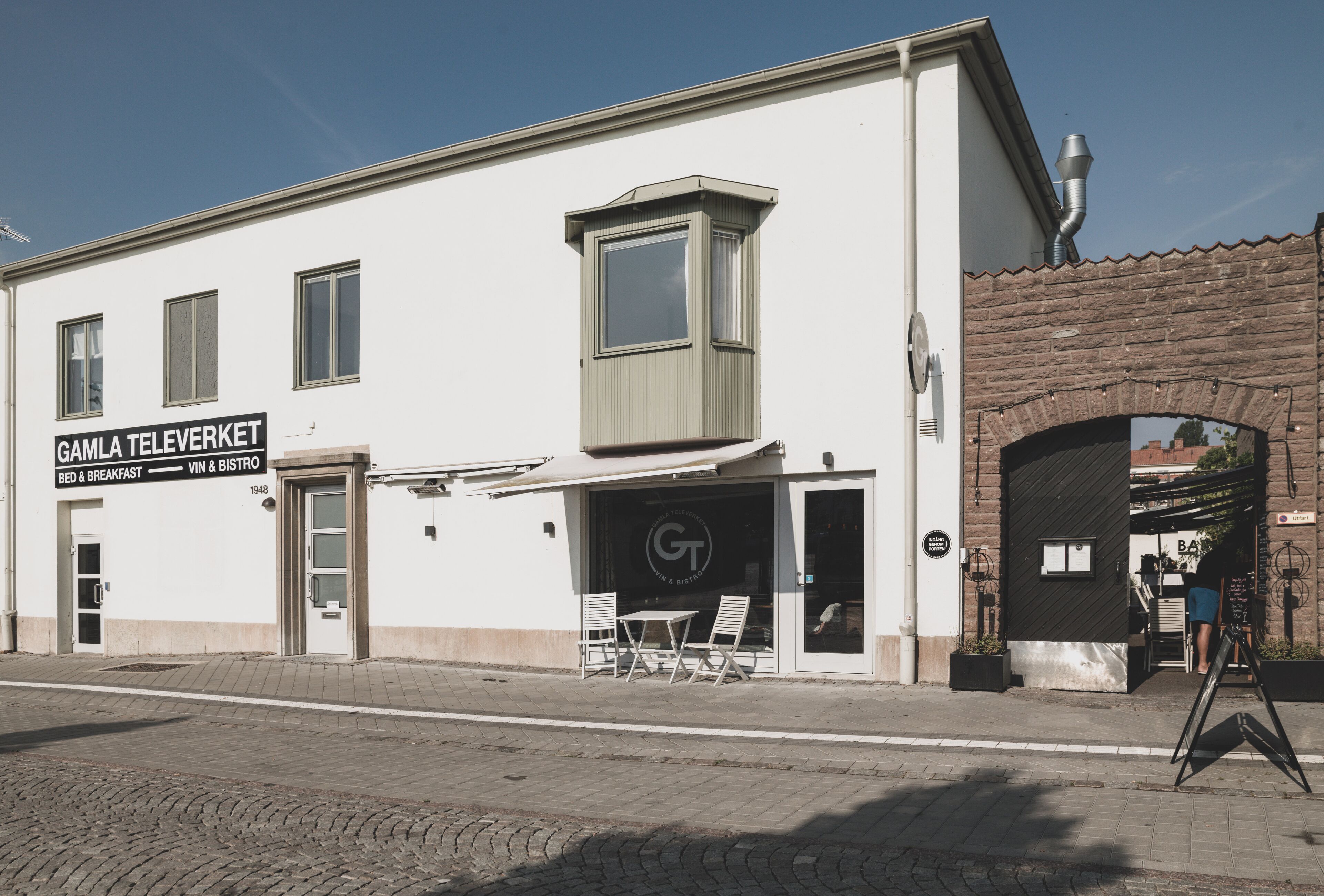 A building with a sign reading "GAMLA TEL EVERKET BED & BREAKFAST" and a small outdoor seating area with tables and chairs.