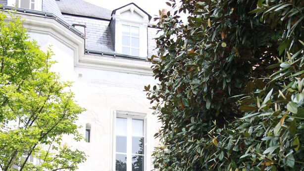 Un bâtiment blanc doté d’une grande entrée en arc de cercle, entouré d’une végétation luxuriante et de plantes en pot.