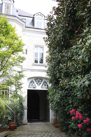 Un bâtiment blanc doté d’une grande entrée en arc de cercle, entouré d’une végétation luxuriante et de plantes en pot.