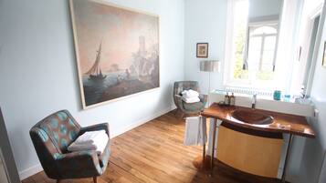 A room with a wooden floor, a large painting of a sailboat, a chair with a towel, a sink, and a window with a view.