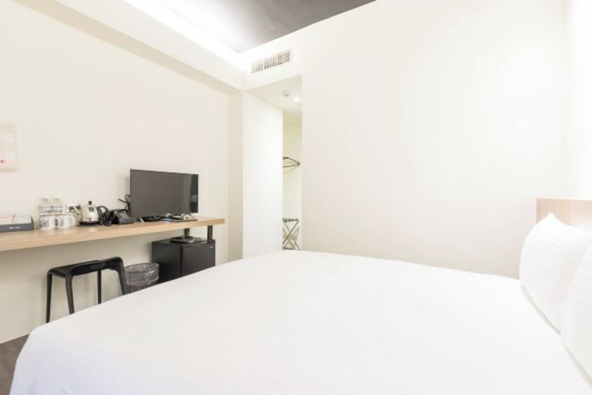 A hotel room with a bed, a TV on a shelf, a small desk with a kettle, and a chair.