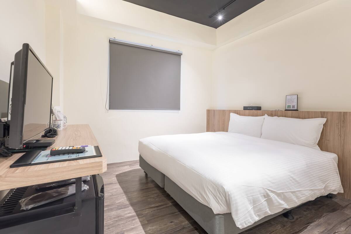 A modern hotel room with a bed, a desk with a computer, and a window with a blind.