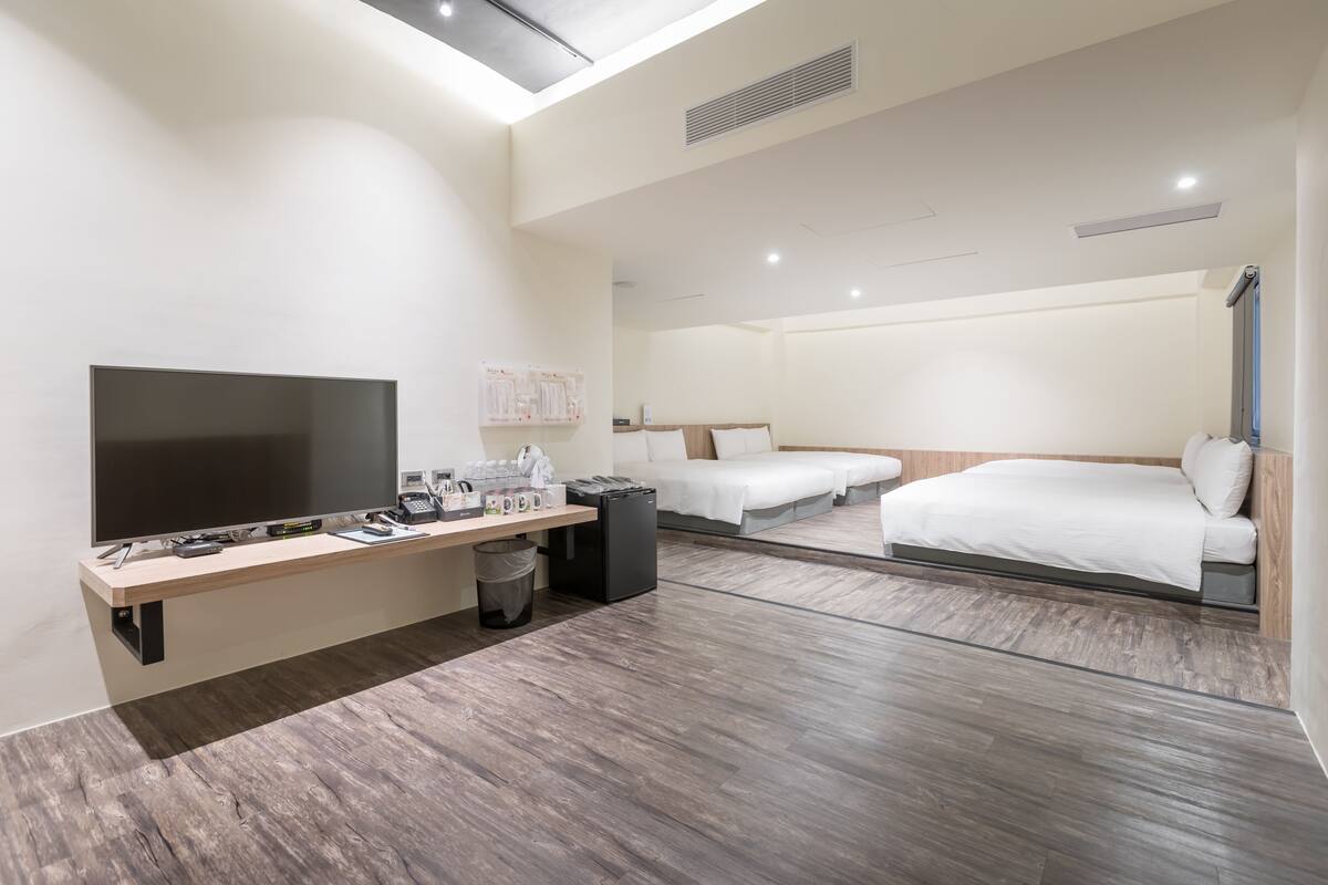 A modern hotel room with a flat-screen TV, a wooden desk, a trash bin, and two beds.