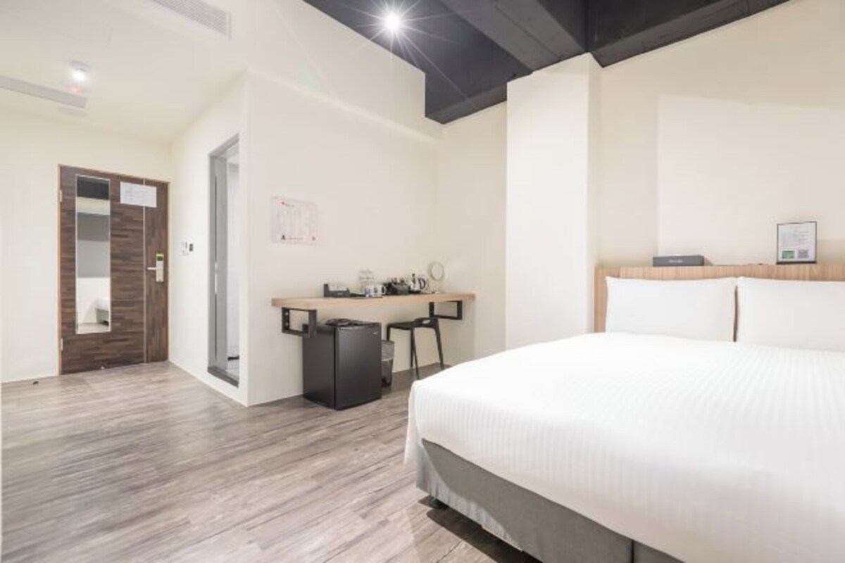A modern hotel room with a large bed, a desk with a chair, and a small trash bin.