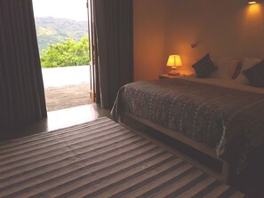 A bedroom with a bed, a nightstand with a lamp, and a view of greenery outside.
