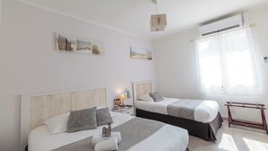 Habitación de hotel con dos camas, una mesita de noche con lámpara, una ventana con cortinas translúcidas y una obra de arte en la pared.