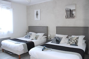 A hotel room with two beds, each with a pillow and a blanket. There is a window on the left side, and artwork is displayed on the walls. - Le Héron Cendré (La Tremblade)