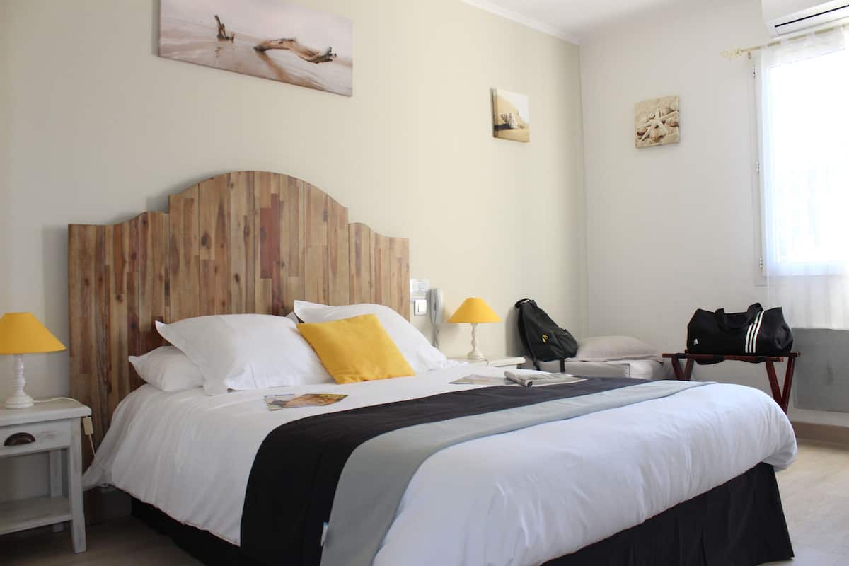 Een hotelkamer met een houten hoofdbord, wit beddengoed, een geel kussen, een zwarte tas op een nachtkastje en kunstwerken aan de muren.
