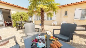 Un área de comedor al aire libre con sillas de mimbre, una mesa dispuesta con croissants, fruta y bebidas, y una palmera de fondo.