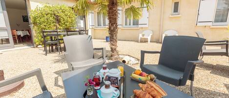 An outdoor dining area with wicker chairs, a table set with croissants, fruit, and beverages, and a palm tree in the background.