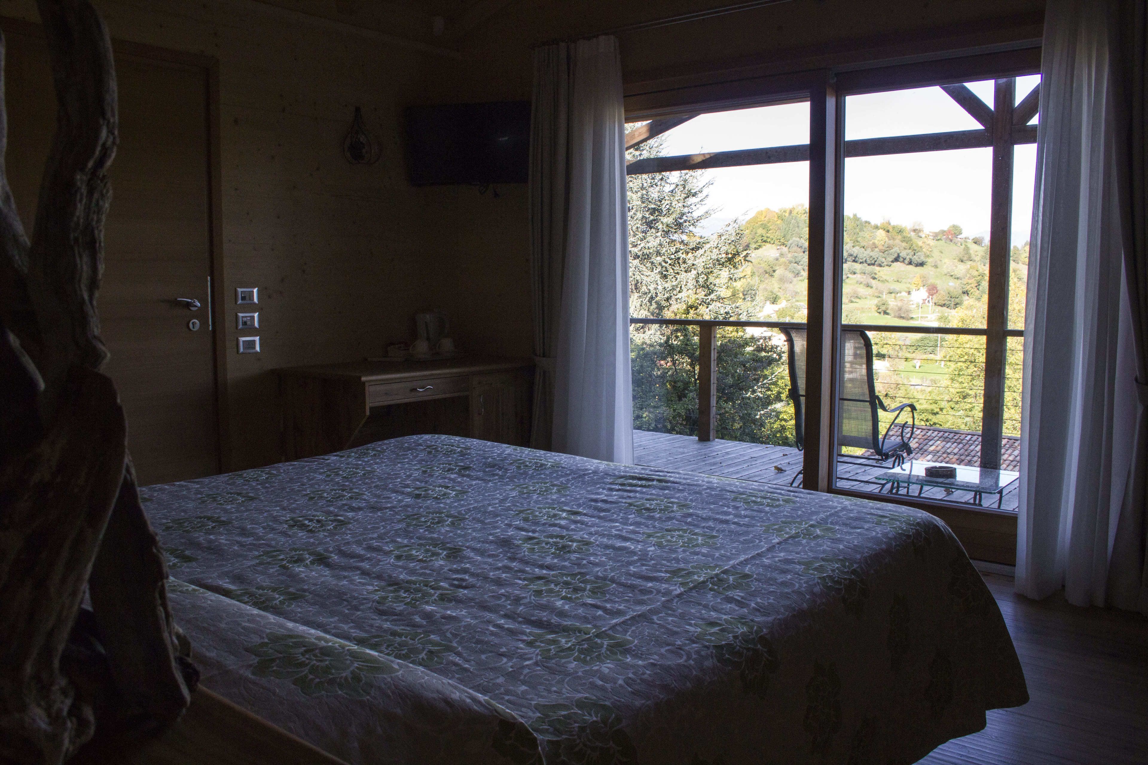Une chambre avec un grand lit, une commode en bois, un téléviseur fixé au mur et un balcon offrant une vue sur la campagne.
