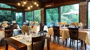 Un restaurante con varias mesas dispuestas para comer, amplias ventanas que ofrecen vistas a zonas verdes y un techo de madera con luces colgantes.