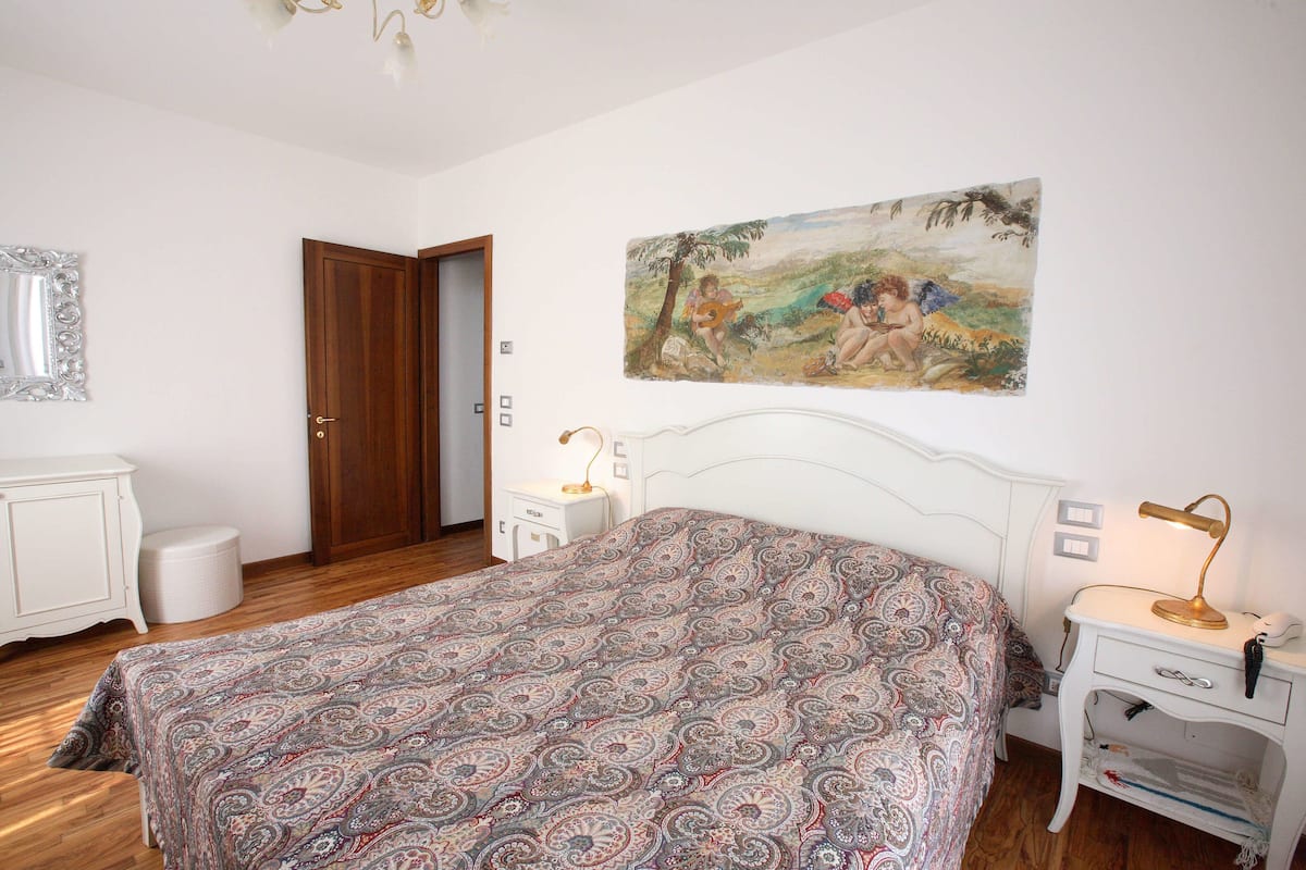 A bedroom with a patterned bedspread, a white dresser, a nightstand with a lamp, and a painting on the wall.