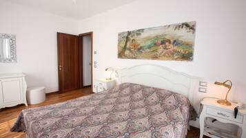 A bedroom with a patterned bedspread, a white dresser, a nightstand with a lamp, and a painting on the wall.