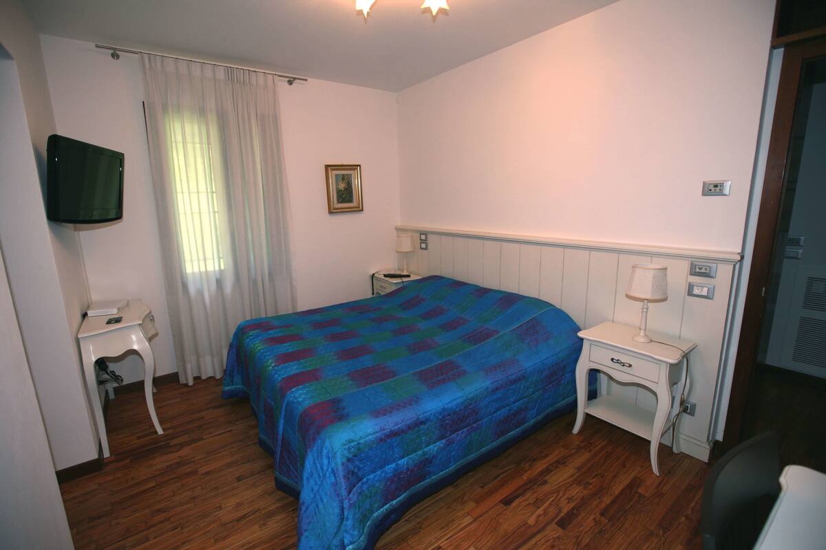 A bedroom with a bed, a bedside table, a lamp, a TV mounted on the wall, and a window with curtains.