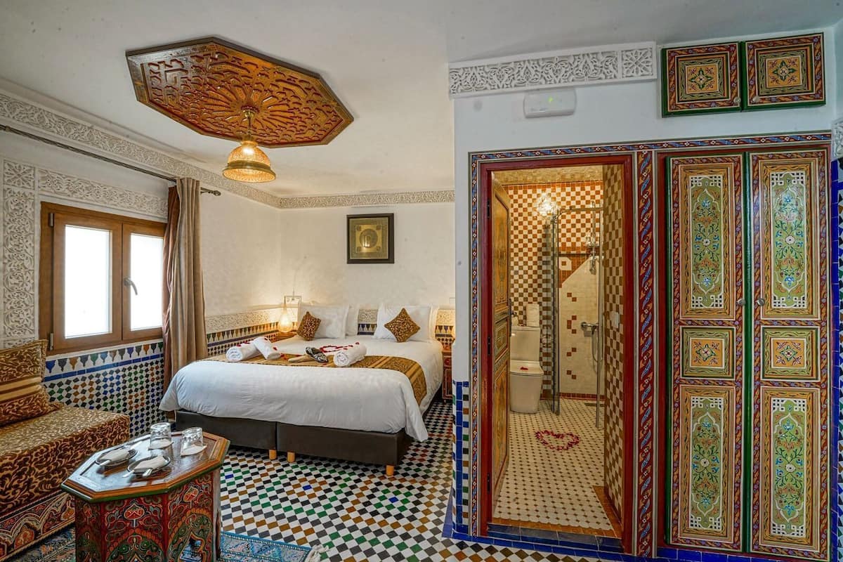 Una habitación tradicional marroquí con suelo de cuadros, una cama con sábanas blancas, un sofá estampado y una lámpara decorativa en el techo.