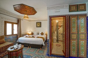 Quarto marroquino tradicional com chão quadriculado, cama com roupa de cama branca, sofá estampado e luminária decorativa no teto.