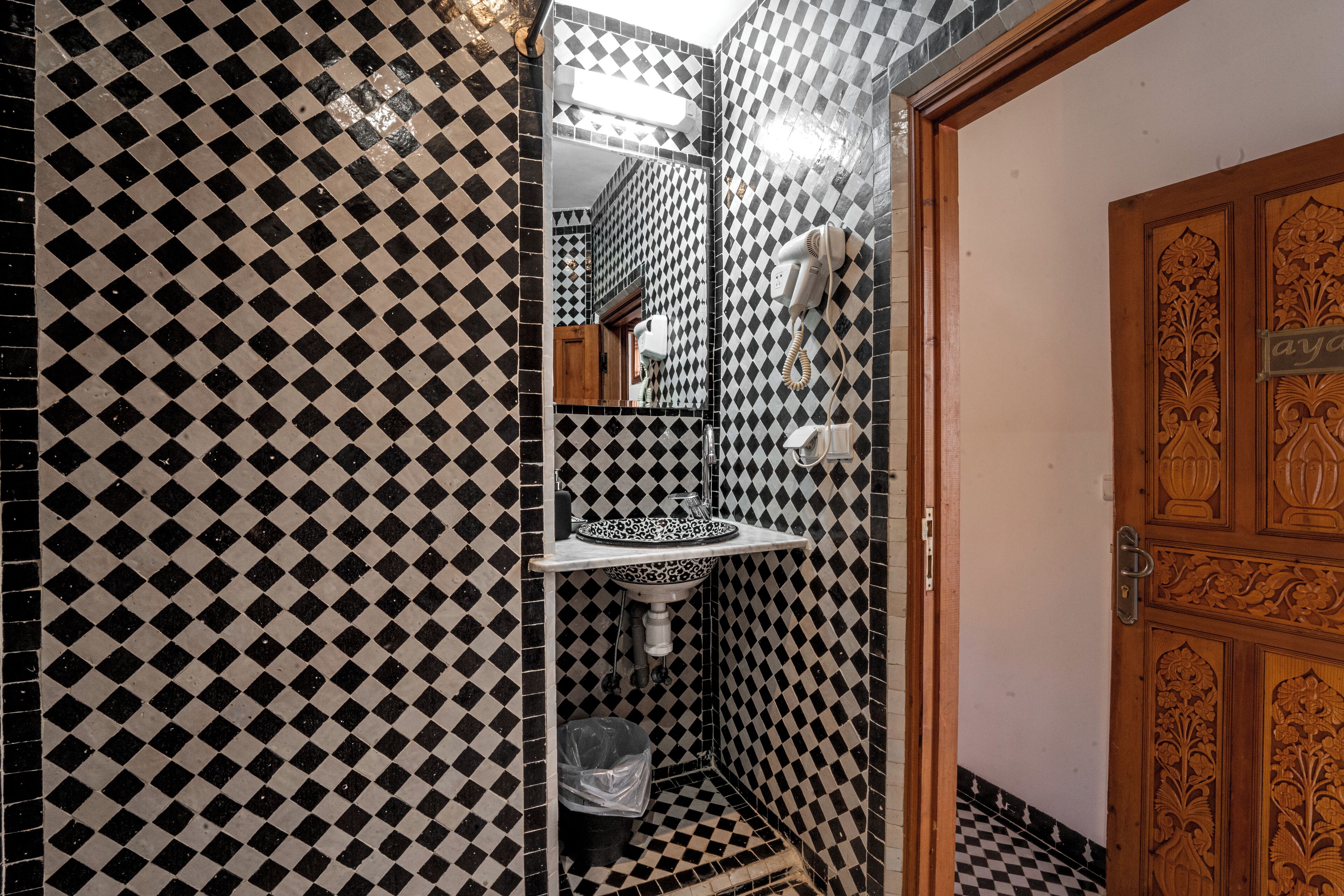 A bathroom with a black and white checkered tile pattern, a white pedestal sink, and a wooden door with intricate carvings.