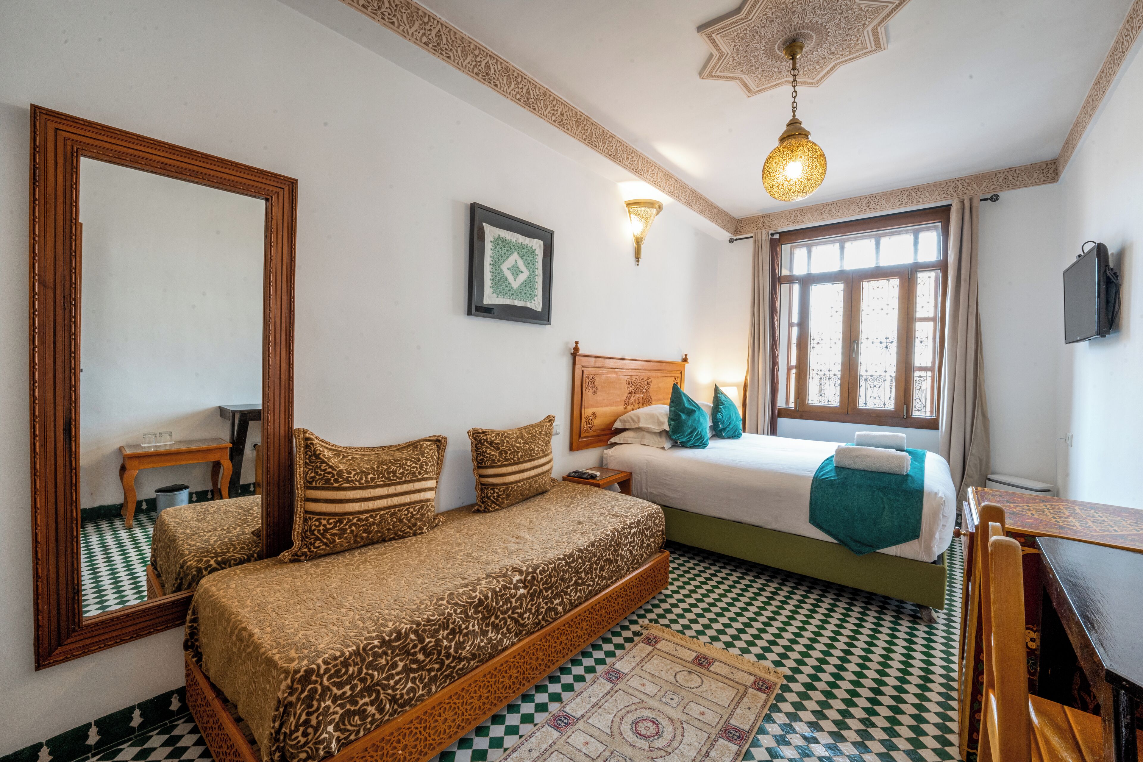A bedroom with a checkered floor, a bed with patterned bedding, a wooden headboard, a bedside table, a mirror, a wall-mounted light, and a window with curtains.
