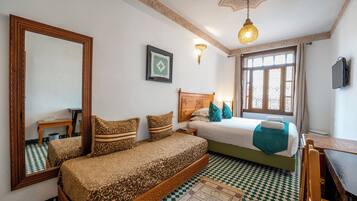 A bedroom with a checkered floor, a bed with patterned bedding, a wooden headboard, a bedside table, a mirror, a wall-mounted light, and a window with curtains.