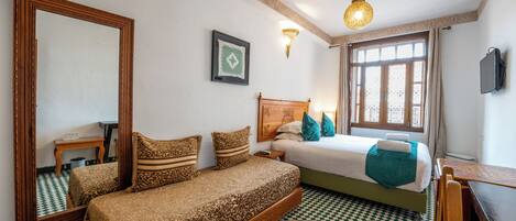 A bedroom with a checkered floor, a bed with patterned bedding, a wooden headboard, a bedside table, a mirror, a wall-mounted light, and a window with curtains.