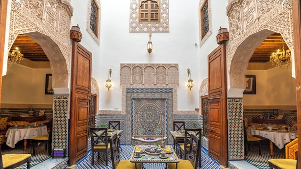 Un interior tradicional marroquí con arcos ornamentales, suelos de azulejos con diseños y una zona de comedor central con mesas y sillas.