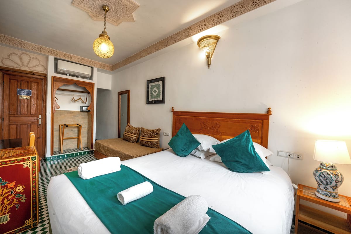 Una cama bien tendida con colcha verde y sábanas blancas, una mesita de noche de madera con una lámpara y un aire acondicionado instalado en la pared.