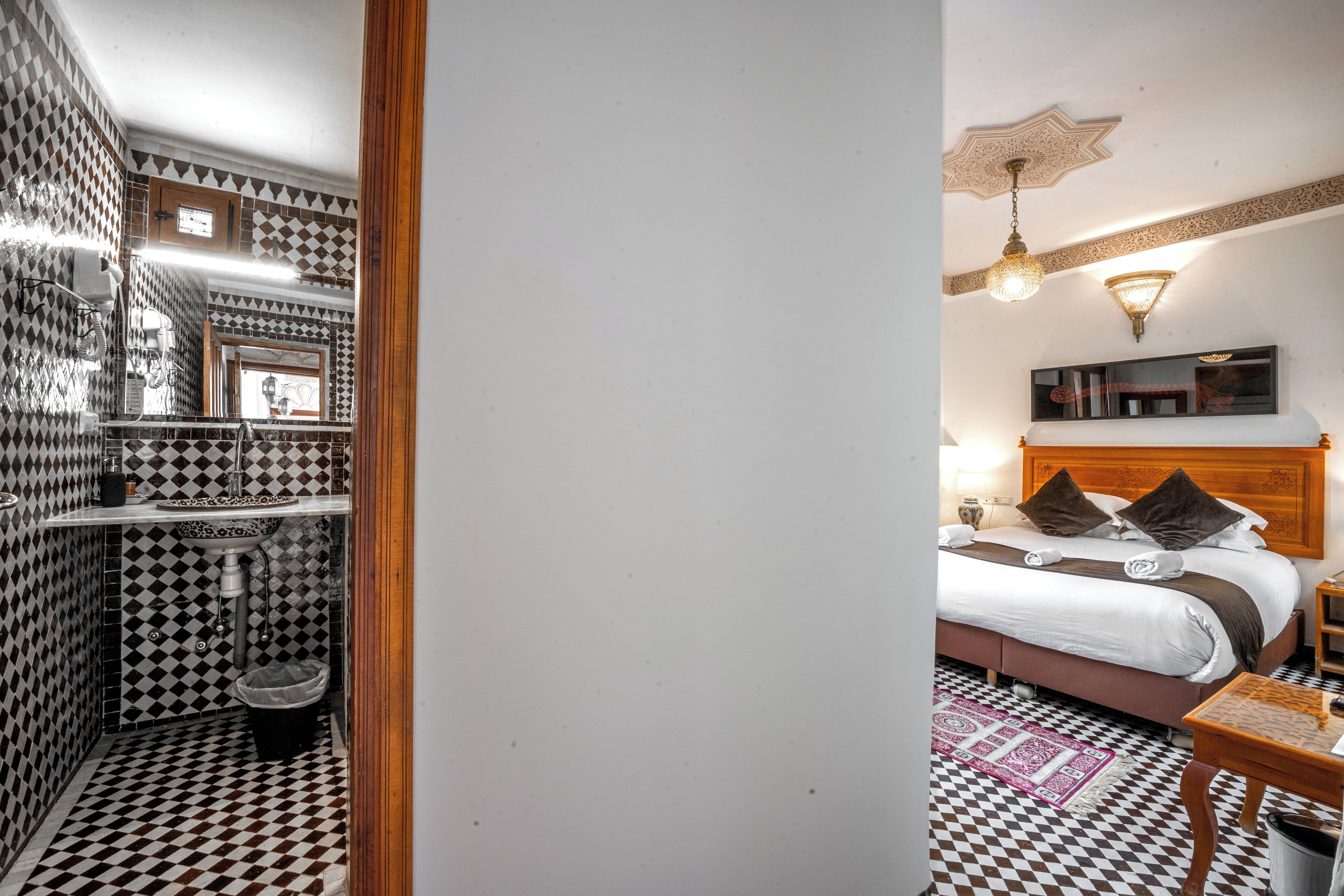 A hotel room with a checkered floor, a bed with a wooden headboard, a nightstand, a bedside table, and a bathroom with a sink and mirror.
