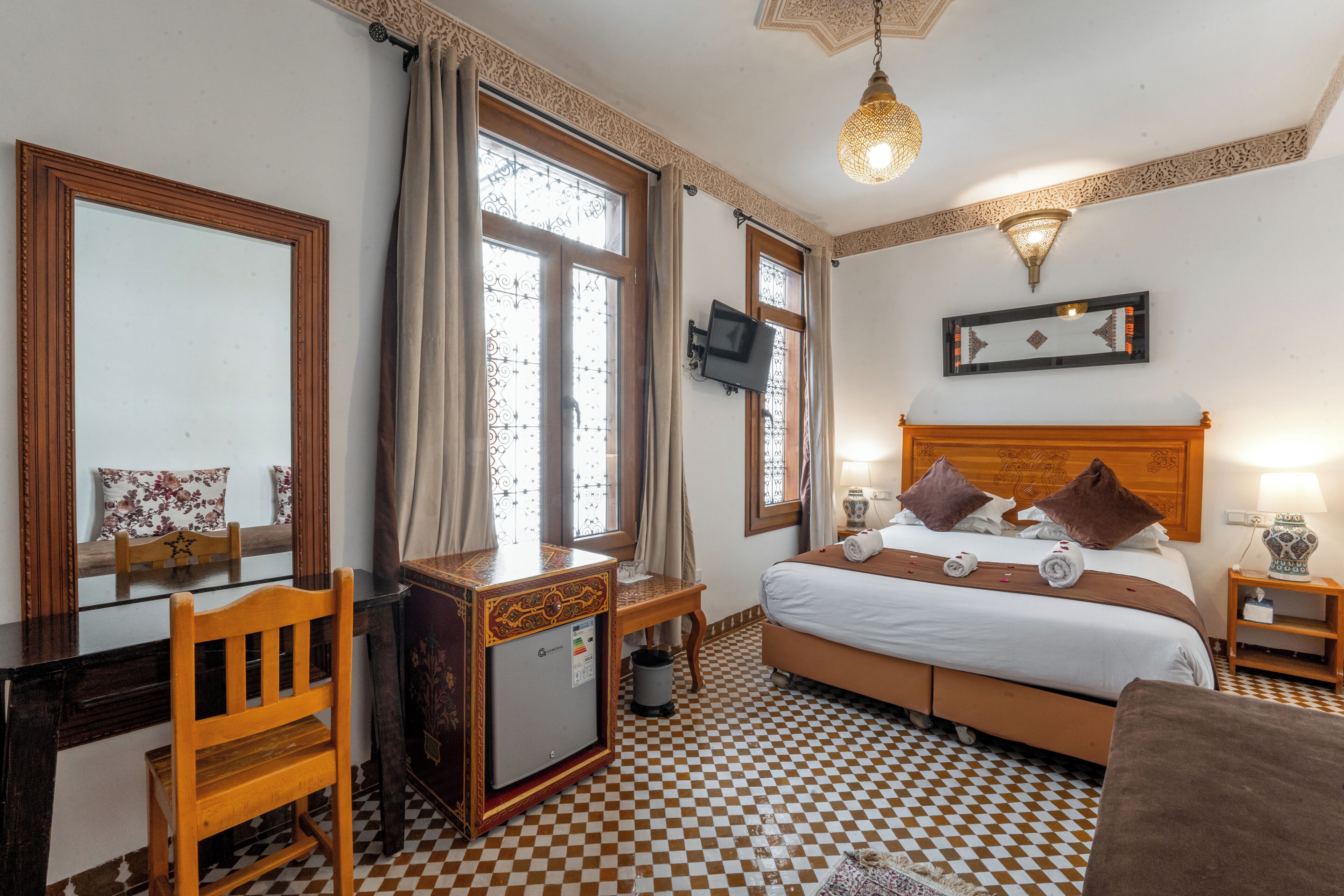A hotel room with a checkered floor, a bed with pillows, a desk with a chair, a television, and a small table with a lamp.