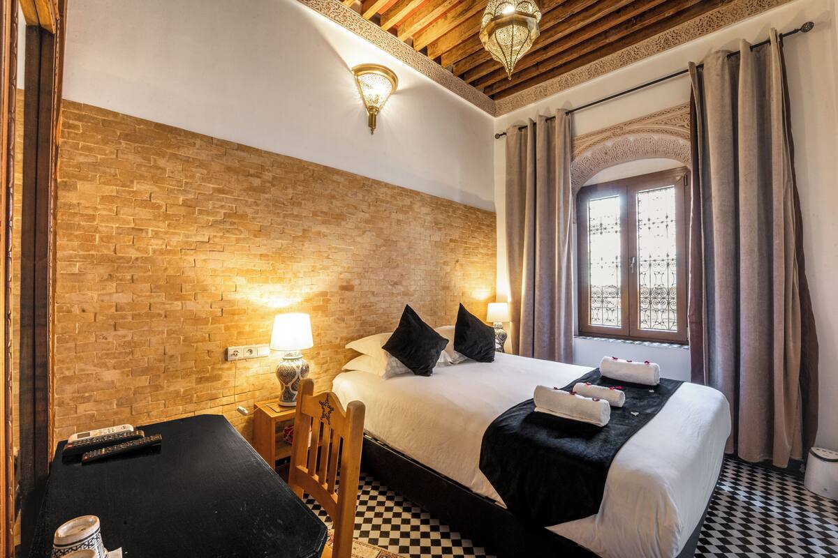 Habitación de hotel con pared de ladrillo, cama con ropa blanca, suelo a cuadros, un escritorio de madera y una ventana con cortinas.