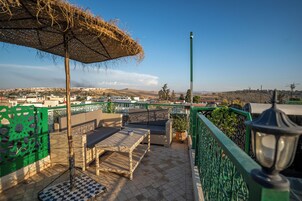 Um terraço com móveis de vime, um guarda-sol de palha e vista para uma cidade ao longe.