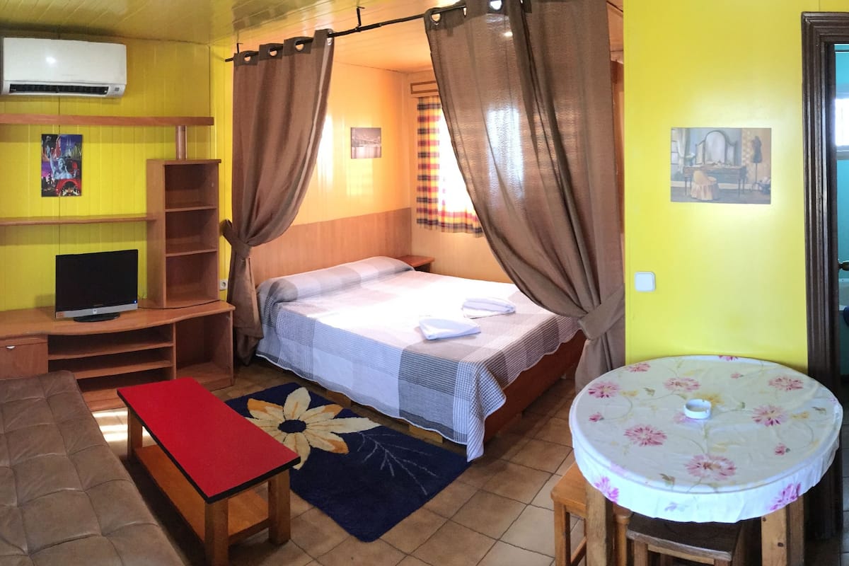 Una habitación con cama, una mesita, televisor y una zona de cocina.