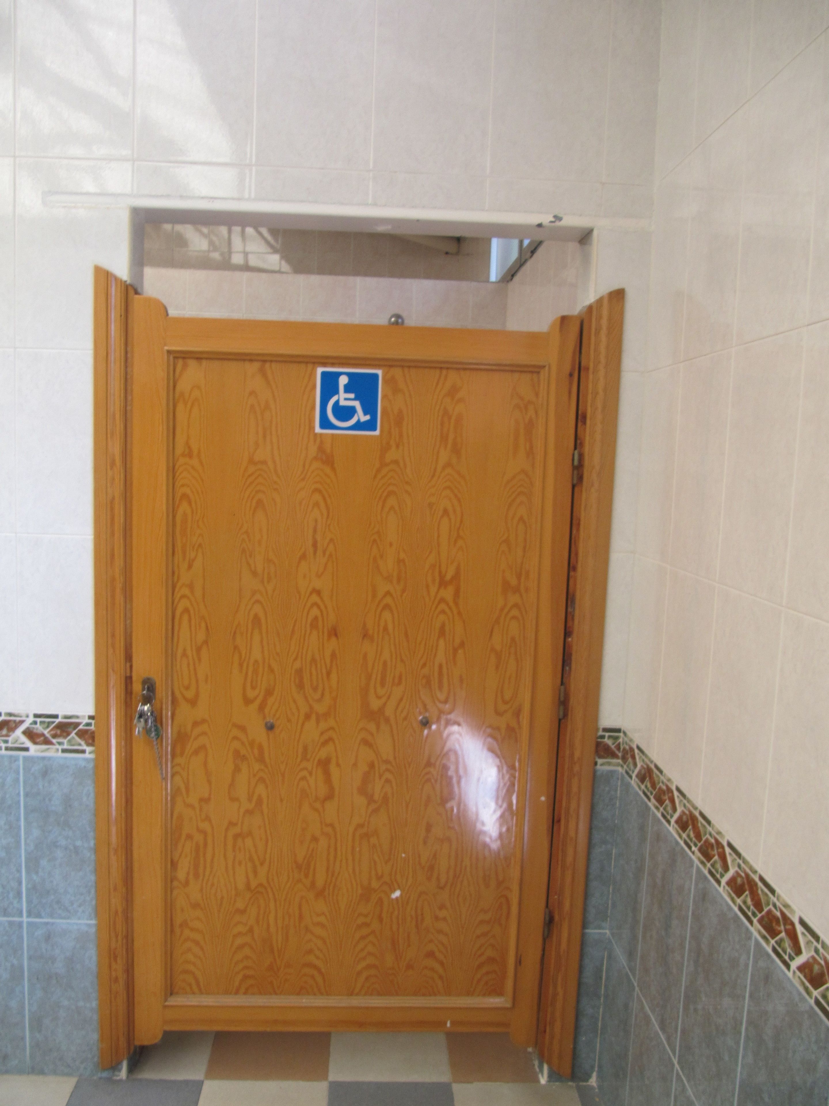 A wooden door with a wheelchair accessibility sign, a tiled floor, and a corner with a decorative tile border.