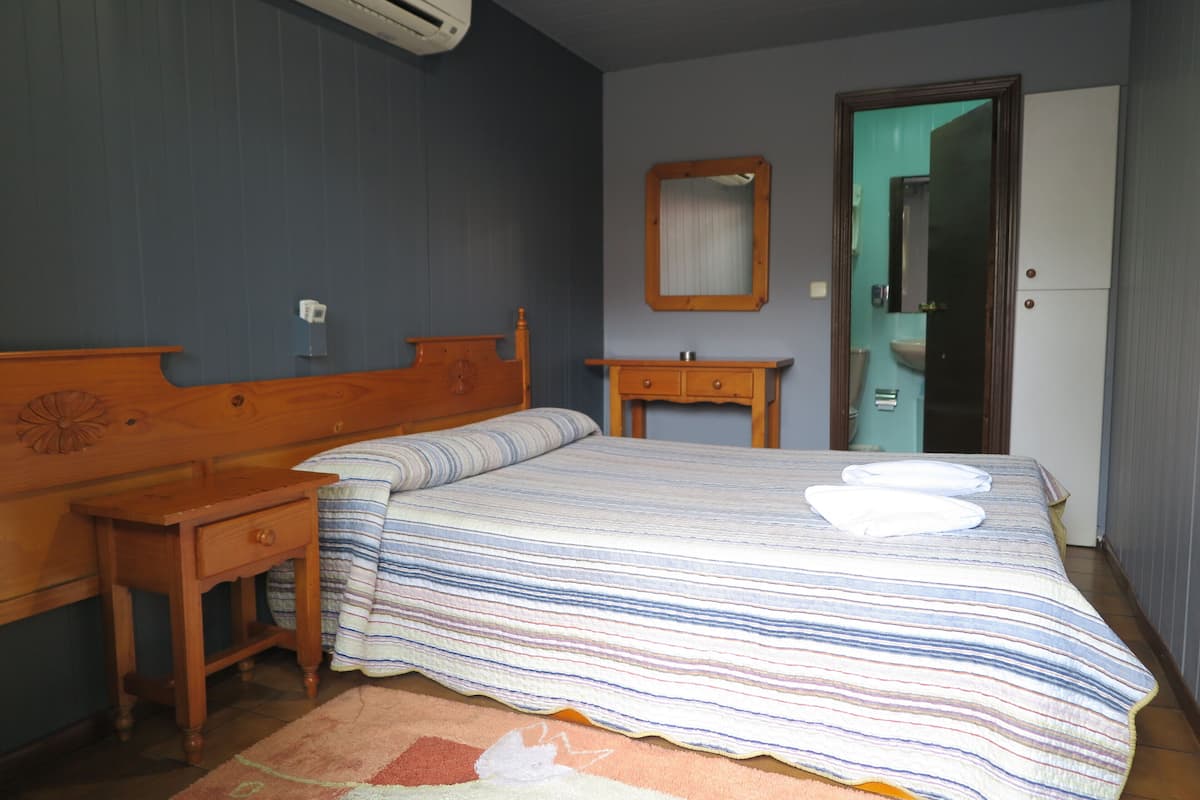 Un dormitorio con cama, mesita de noche de madera, cómoda y un baño visible a través de una puerta abierta.