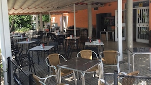 Un café al aire libre con mesas y sillas, una zona cubierta para sentarse y un edificio al fondo.