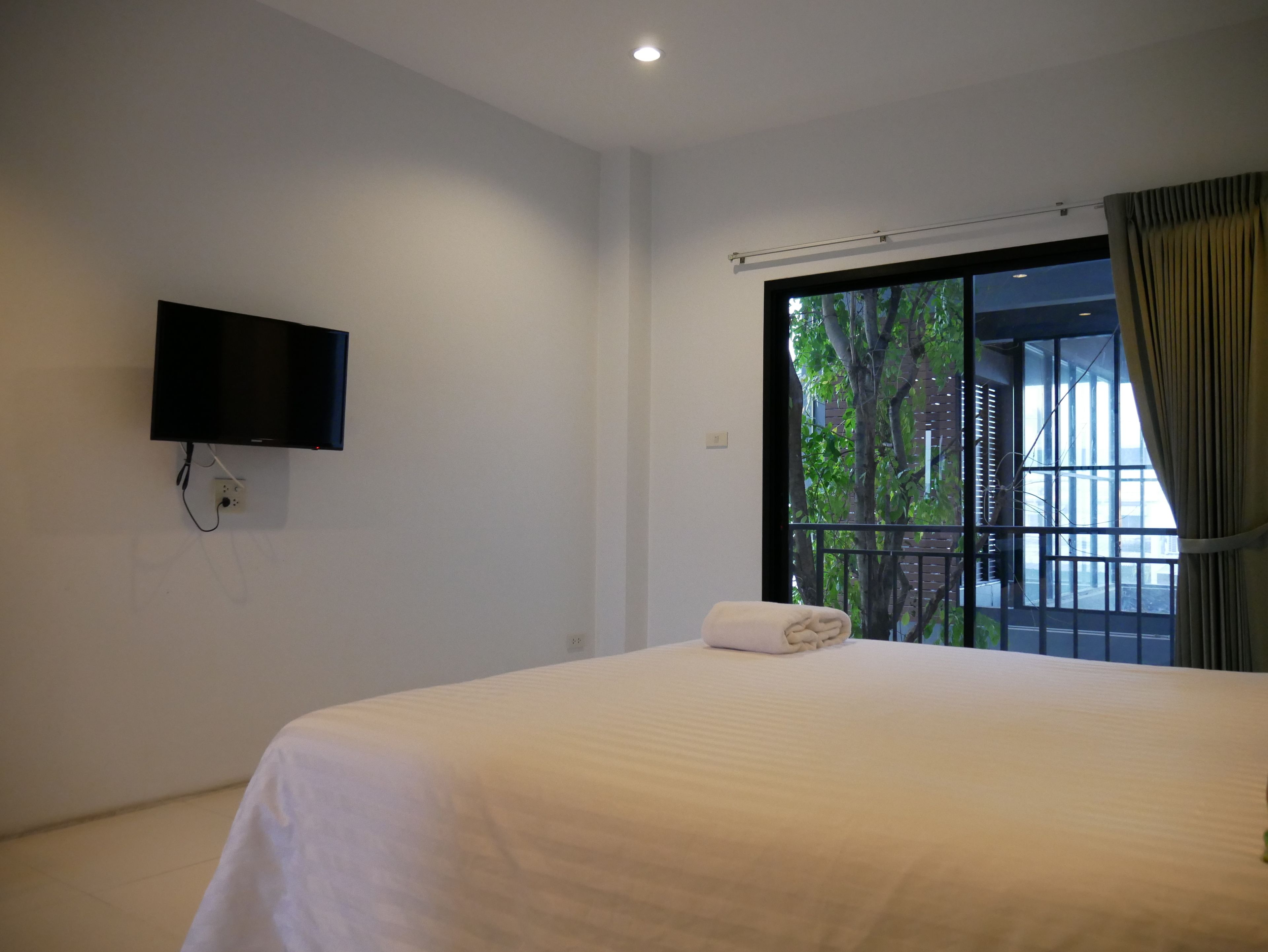 A hotel room with a bed, a flat-screen TV mounted on the wall, and a window with a view of trees.