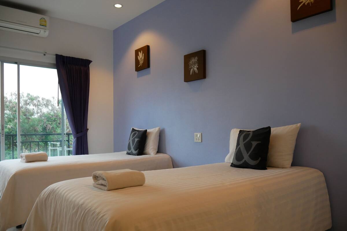 Ein Hotelzimmer mit zwei Betten, einem fensterbelaubten Fenster, einer Klimaanlage und zwei Wandbildern.
