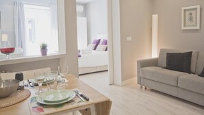 Una sala moderna con un sofá, una mesa de comedor puesta con platos, vasos y cubiertos, y vista a otra habitación con una cama y un ventanal.